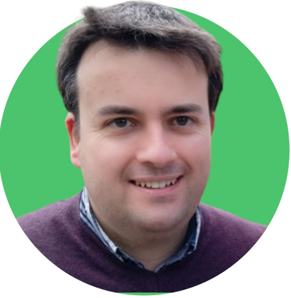 La photo montre un homme au teint clair avec des cheveux bruns courts et soignés. Il porte un pull violet sur une chemise à col boutonné à carreaux bleus et blancs. L'homme sourit doucement, donnant une impression de cordialité et d'accessibilité. Le fond de l'image est un vert uni, ce qui met en avant le sujet sans distraction.